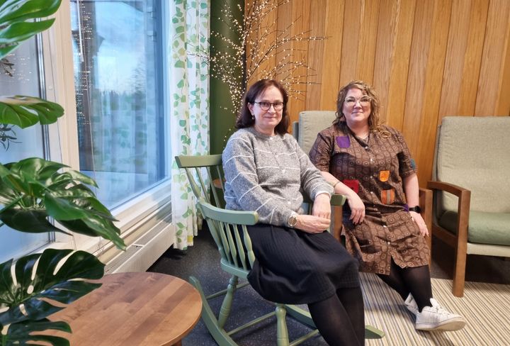 Valokuva kahdesta naisesta istumassa viihtyisässä sisätilassa, jossa on puupaneelit ja viherkasveja.