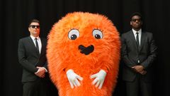 Oranssi, karvainen maskottihahmo seisoo kahden mustiin pukeutuneen henkivartijan välissä