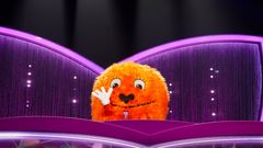 Oranssi, karvainen maskottihahmo vilkuttelee Masked Singer -ohjelman tuomaripöydän takaa