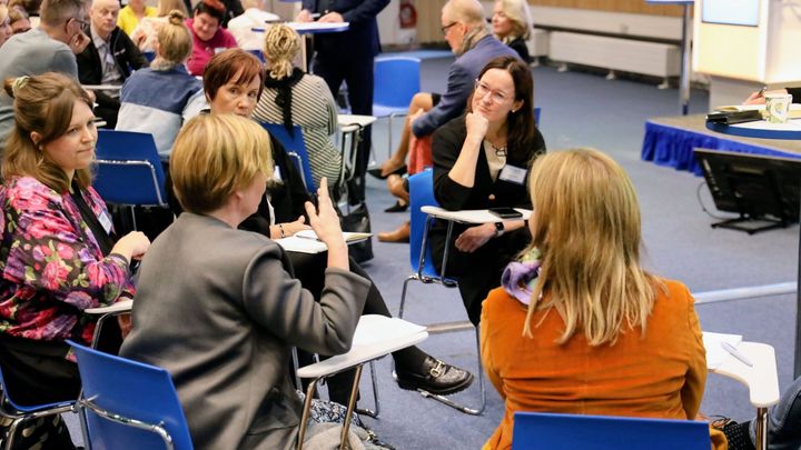 Osallistujat keskustelevat seminaarissa istuen sinisillä tuoleilla.