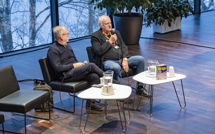 Seppo Jokinen puhuu mikrofoniin istuen lavalla toisen miehen kanssa, edessä pienet pöydät vesilaseineen ja kirja.