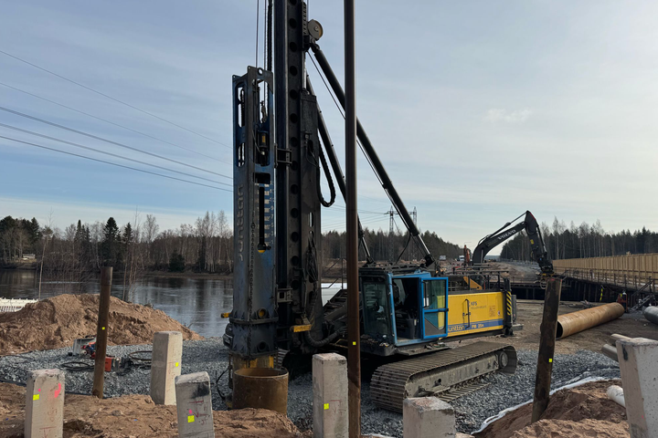 Kuva rakennustyömaalta, jossa suuri työkone paaluttaa, taustalla joki ja metsää.