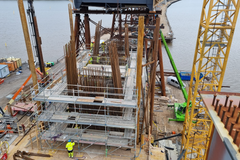 Rakennustyömaa meren äärellä, jossa keskeneräinen teräsbetonirakenne, jalusta, kohoaa rakennustelineiden ja rautojen keskeltä. Taustalla näkyy vesi ja pilareita.