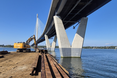 Rakennustyömaa Kruunuvuorensillalla: kaivinkone työskentelee purkamassa työsiltaa veden äärellä. Taustalla kohoaa massiivinen betonisilta pilvettömänä syyspäivänä.