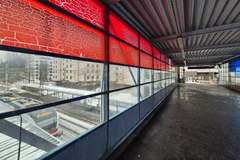 Alt-teksti: Kannelmäen juna-aseman ylikäytäväsilta, jossa on punaisia ja sinisiä lasipaneeleita, näkyy sateisena päivänä. Rakennuksia ja junan vaunu näkyvät taustalla.