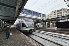 Asemaalue, jossa juna pysähtyneenä laiturilla, taustalla näkyy lasinen kävelysilta ja kerrostaloja. Henkilö kävelee poispäin junasta.