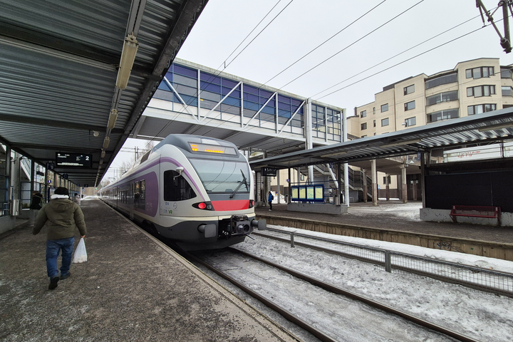 Asemaalue, jossa juna pysähtyneenä laiturilla, taustalla näkyy lasinen kävelysilta ja kerrostaloja. Henkilö kävelee poispäin junasta.