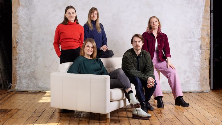 Viisi ihmistä poseeraa sisätiloissa puisella lattialla valkoisen sohvan ympärillä.