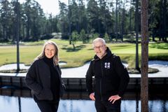 Kaksi henkilöä, nainen ja mies, seisoo ulkona golfviheriön ja veden äärellä.