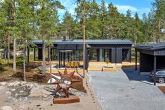 Kuvassa yksikerroksinen, tumma, puupintainen moderni omakotitalo, joka on sijoittuneena mäntyjä kasvavan metsän reunaan. Talon läheisyydessä on kalliota ja kanervikkoa. Kuvan etualalla on omakotitalon etupiha, joka on laattapäällysteinen. Kuvan oikeassa laidassa on autokatos, jossa parkkeerattuna farmarimallinen BMW. Aivan kuvan etualalla on tähden muotoinen veistos, jonka materiaali on ruostunutta rautaa.