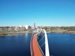 Silta, joka muistuttaa linnunsiipiä, ylittää Oulujokisuiston kirkkaan sinisen veden. Taustalla näkyy kaupunkimaisemaa.