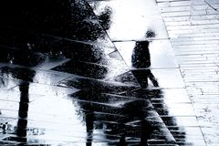 Yksinäinen nuori kävelemässä kaupungilla sateessa, heijastuu vesilätäköstä.