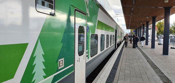 Matkustajajuna asemalaiturilla kuvan vasemmalla laidalla, pitkä junan kylki näkyvissä. Kauempana matkustajia asemalaiturilla.