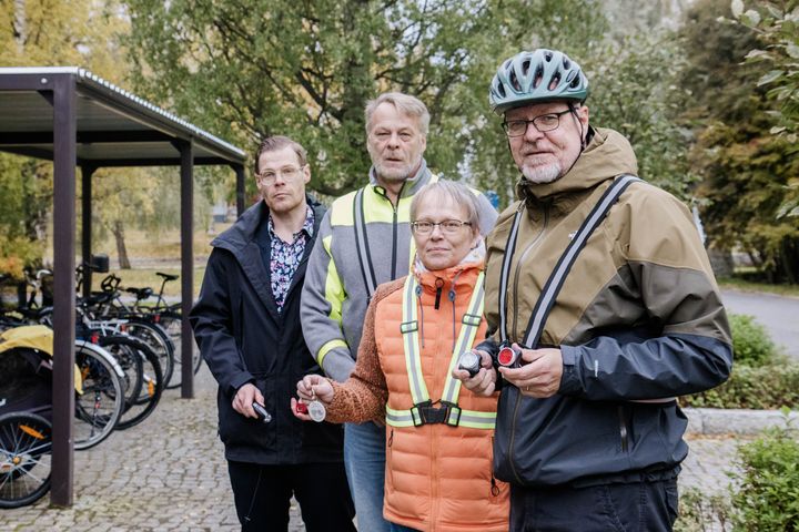 Kolme miestä ja yksi nainen seisoo ulkona pyörätelineiden edustalla. Yhdellä miehistä on päässään pyöräilykypärä, kaikilla on asussaan erilaisia heijastimia.