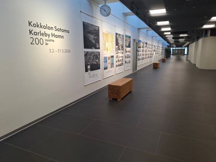 Näkymä leveälle käytävälle, jonka valkoisella seinällä useita valokuvia.