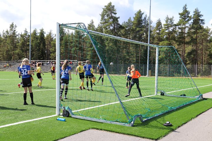 En fotbollsmatch där kvinnor spelar på en konstgräsplan, med skog i bakgrunden.