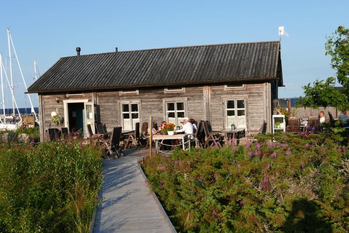 Harmaantuneella puulla verhoiltu kahvilarakennus, jonka edustalla istuu asiakkaita ulkona. Aurinko paistaa. Rakennukselle vie puinen kävelytie