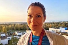 Rintakuva Anu Arosesta, joka on pukeutunut siniseen paitaan ja valkoiseen takkiin. Taustalla rakennuksia, metsää ja taivasta.