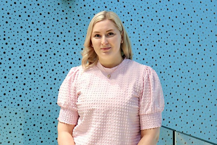 Puolivartalokuva Laura Oksasta, jolla on yllään vaaleanpunainen puhvihihainen paita. Taustalla on turkoosi seinä, jossa on pieniä mustia pisteitä.