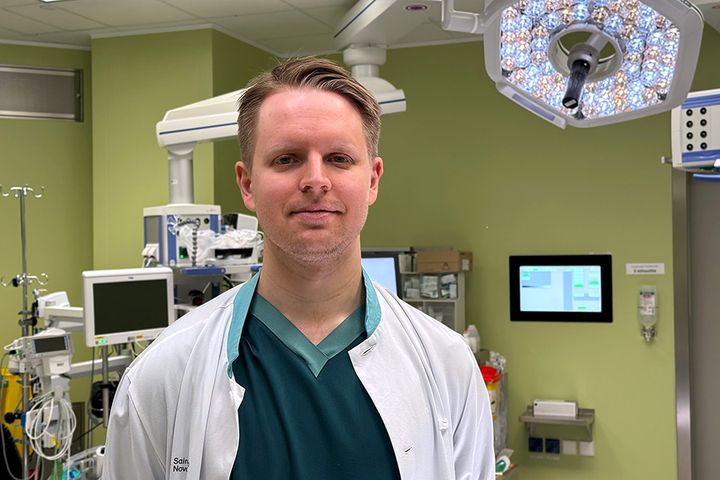 Puolivartalokuva Lauri Nyrhistä, jolla on yllään tummanvihreä paita ja valkoinen lääkärintakki. Ympäröivässä huoneessa on vaaleanvihreät seinät ja erilaisia sairaalalaitteita.