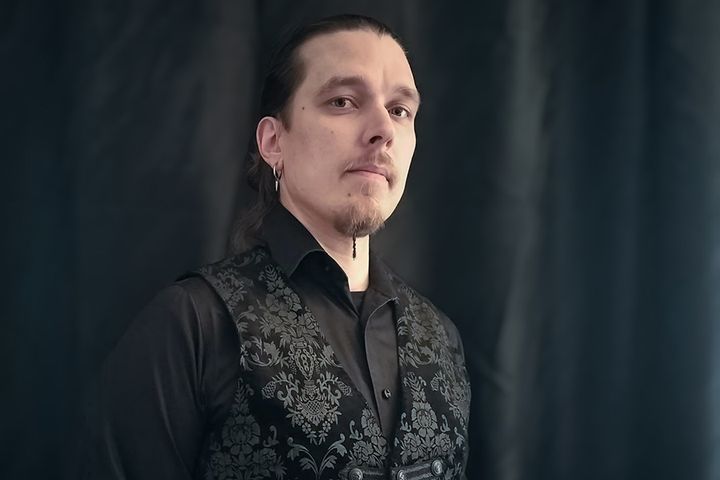 Henkilö mustassa paidassa ja kuvioidussa liivissä, katsoo kameraan. Tausta on tumma.