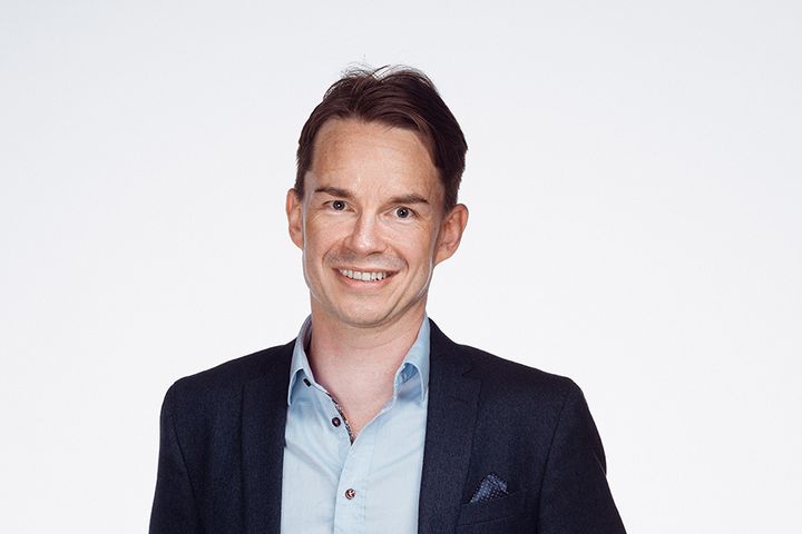 Hymyilevä henkilö, jolla on tummat hiukset, vaalea kauluspaita ja tumma pikkutakki.