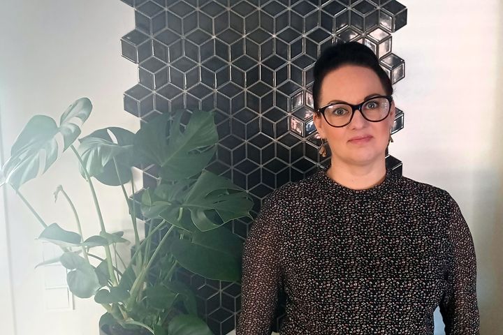 Henkilö seisoo seinän edessä, jossa on musta geometrinen kuvio. Vieressä on viherkasvi.