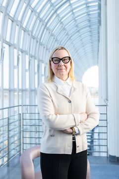 Kuvassa on nainen, joka seisoo lasikattoisella käytävällä, kädet ristissä. Hänellä on yllään vaalea pusero ja mustasankaiset silmälasit.