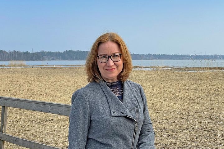 Nainen seisoo ulkona hymyillen, taustalla on järvimaisema.