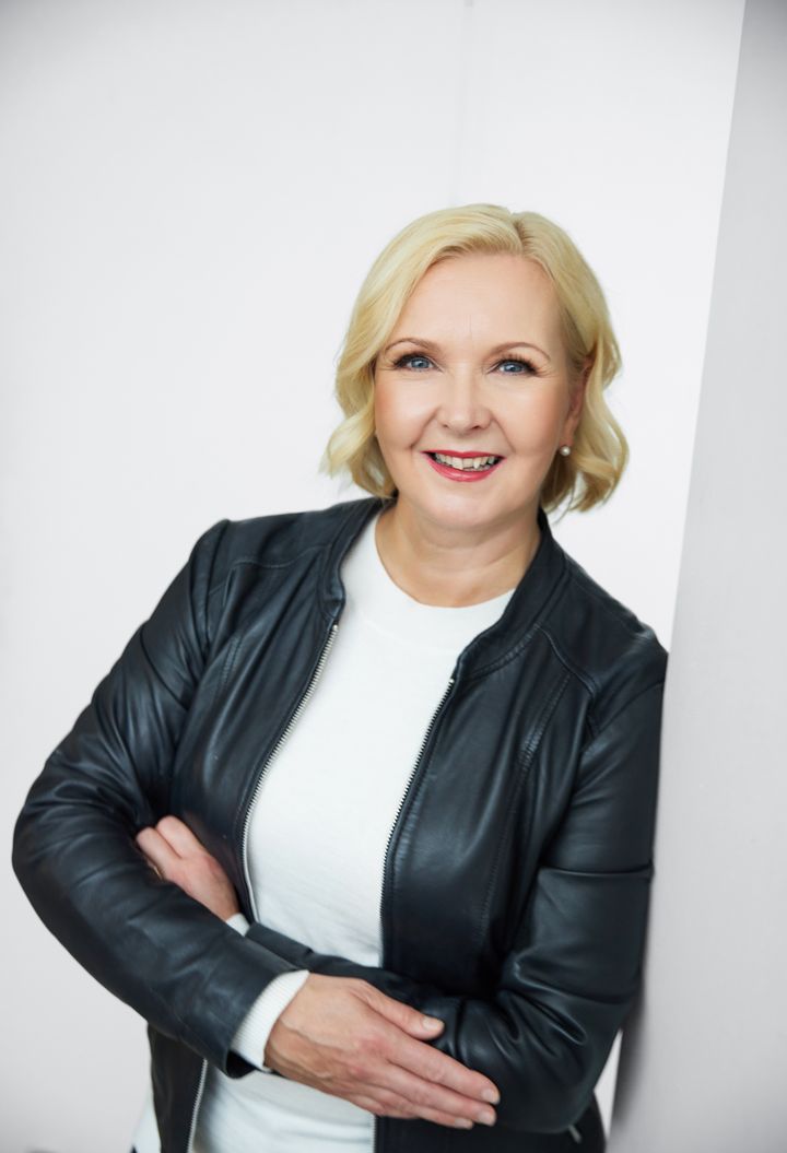 Vaaleahiuksinen nainen nojaa seinään ja katsoo hymyillen kameraan.