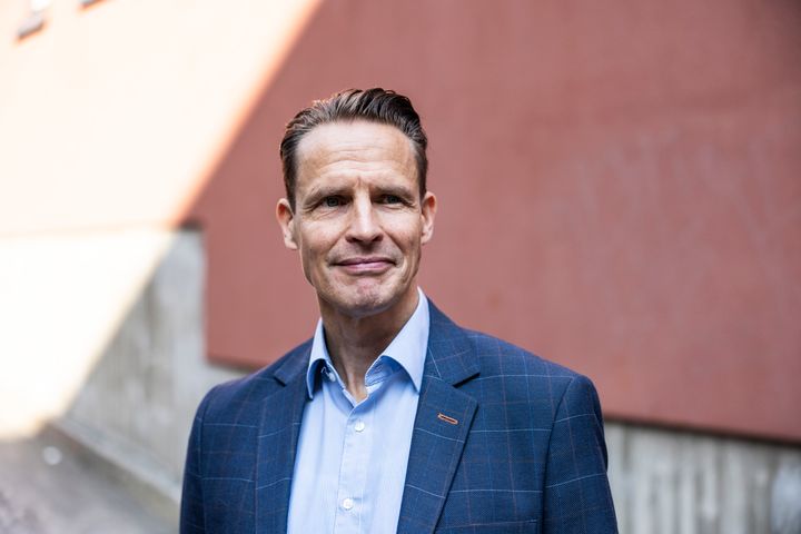 Mies pukeutunut siniseen pukuun ja vaaleansiniseen paitaan seisten ulkona. Tausta on punertava seinä.