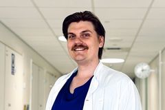 Henkilö valkoisessa lääkärintakissa seisoo käytävällä ja hymyilee.