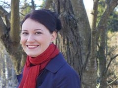 Kuvassa hymyilevä henkilö ulkona puun edessä. Henkilö on pukeutunut punaiseen huiviin ja siniseen takkiin. Taustalla näkyy talvisia puita.