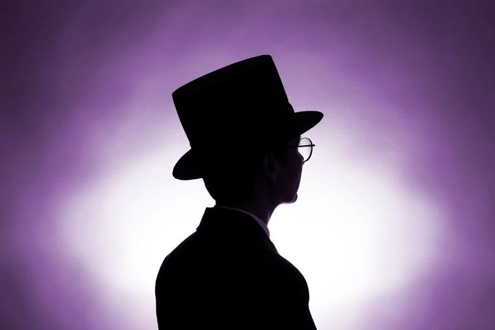 Korkeaa hattua käyttävä henkilö siluetissa violettia taustaa vasten.