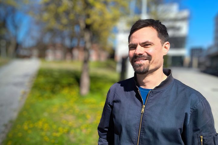 Siniseen takkiin pukeutunut henkilö katsoo hymyillen ohi kamerasta. Taustalla on rakennuksia, nurmikkoa ja puita.