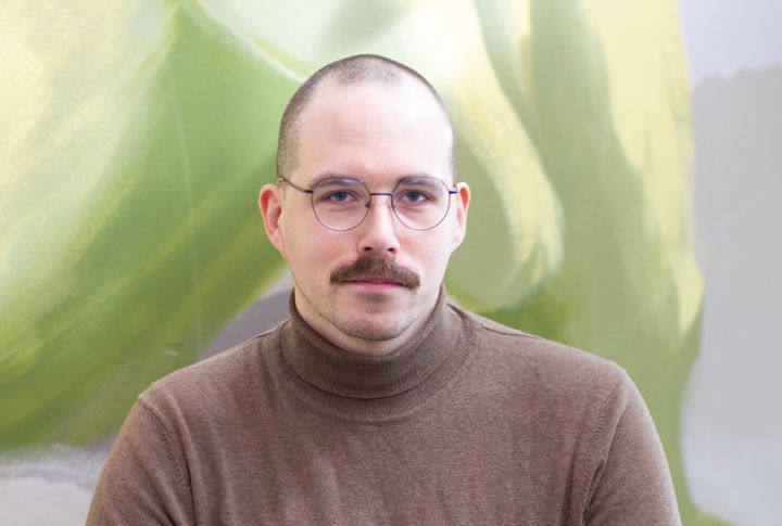 Aikuinen henkilö, jolla on yllään ruskea poolopaita, taustalla vihreitä ja keltaisia sävyjä.