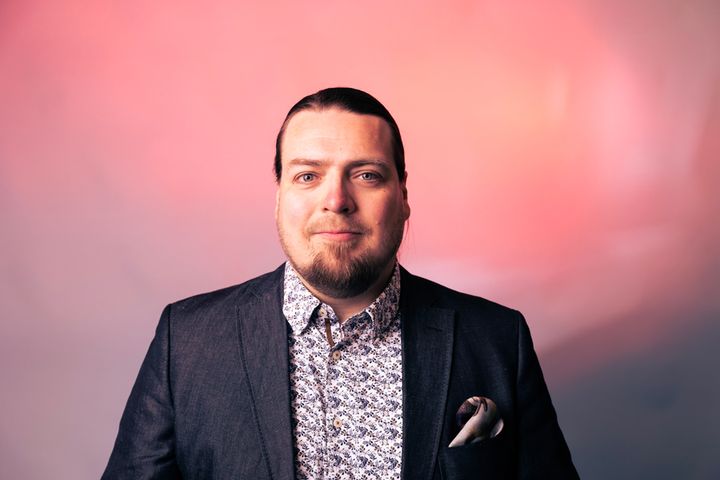Mies pukeutuneena tummaan pukuun ja kuviolliseen paitaan, taustalla vaaleanpunaisen sävyinen pinta.