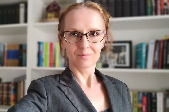 Henkilö seisoo kirjahyllyn edessä ja katsoo kameraan. Hänellä on silmälasit ja pukeutunut pikkutakkiin.