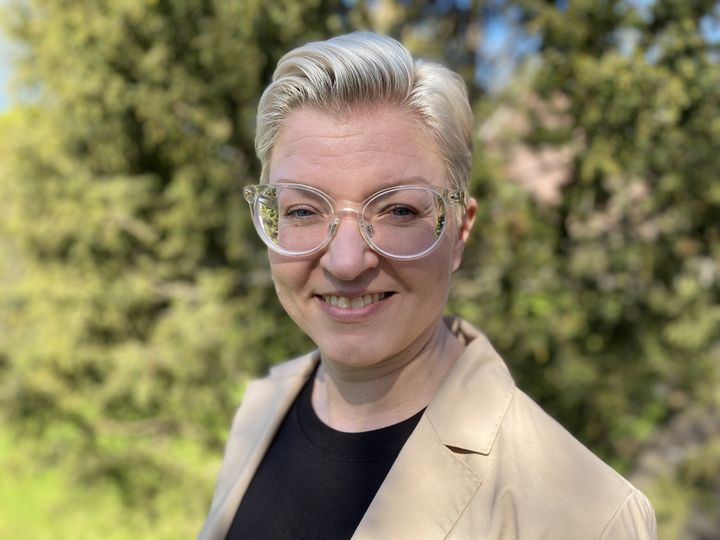 Hymyilevä henkilö ulkona, taustalla vihreää kasvillisuutta. Henkilöllä on vaalea lyhyt tukka, läpinäkyvät silmälasit, musta paita ja vaalea takki.