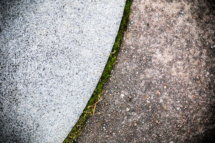 Rajapinta kahden eri tekstuurin välillä: harmaa graniitti ja karkeapintainen asfaltti, joiden välissä ohut sammalrivi.