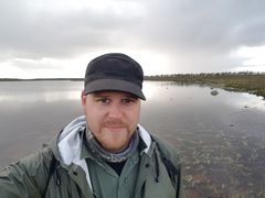 Lauri Jaamala vihreässä sadetakissa syksyisen järvimaiseman edessä.