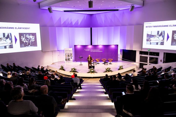 Konferenssisali täynnä kuuntelijoita, esiintyjä puhuu lavalla. Kaksi suurta valkokangasta esittää diaesityksiä.