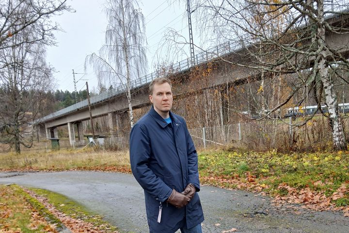 Sinitakkinen mies seisoo ulkona taustallaan betonisilta.