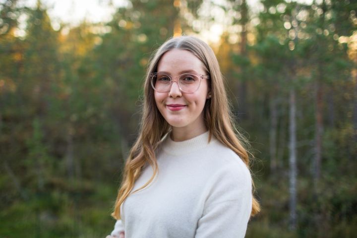 Henkilö seisoo ulkona metsäisessä ympäristössä, yllään vaalea paita ja kasvoillaan hymy.