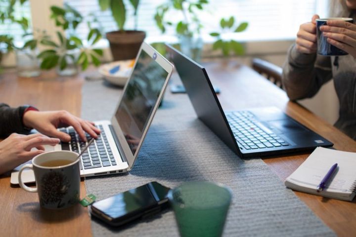 Kaksi henkilöä työskentelee kannettavilla tietokoneilla puupöydän ääressä. Pöydällä on kahvikuppeja, muistilehtiö ja kynä.