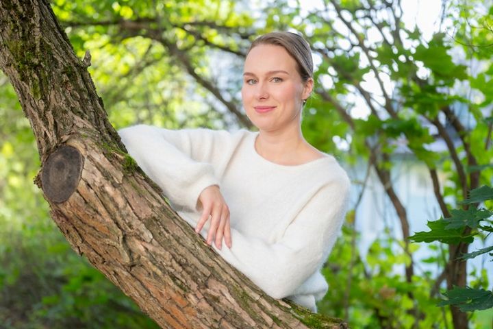 Henkilö valkoisessa neuleessa nojaamassa puun oksalla. Taustalla vihreää lehtimetsää ja hieman vettä näkyvissä.