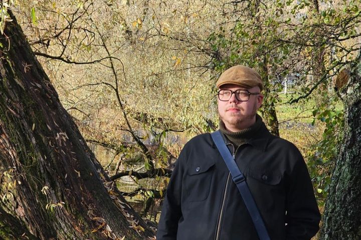 Henkilö seisoo syksyisessä metsässä, jossa on keltaisia ja ruskeita lehtiä. Hänellä on lippalakki ja musta takki yllään.