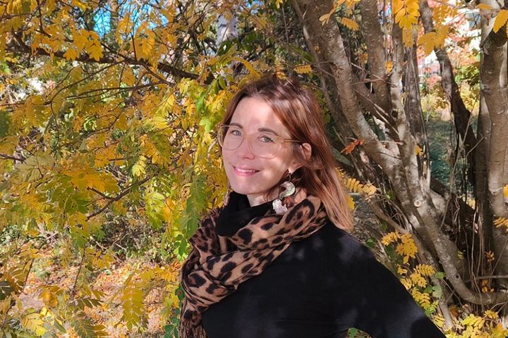 Henkilö seisoo syksyisessä metsässä, värikkäiden lehtien ympäröimänä, ja hymyilee. Hänellä on yllään tumma paita ja leopardikuvioinen huivi.