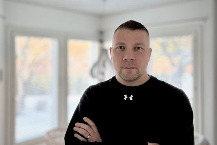 Mies seisoo sisätiloissa rennossa asennossa. Taustalla näkyy ikkunoita.