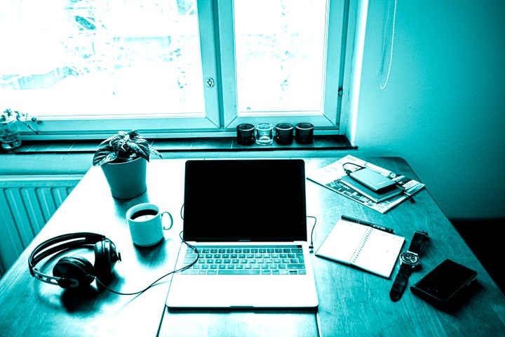 Kannettava tietokone puupöydällä, vieressä kuulokkeet, kahvikuppi, kasvi, muistilehtiö ja kännykkä. Taustalla ikkuna.